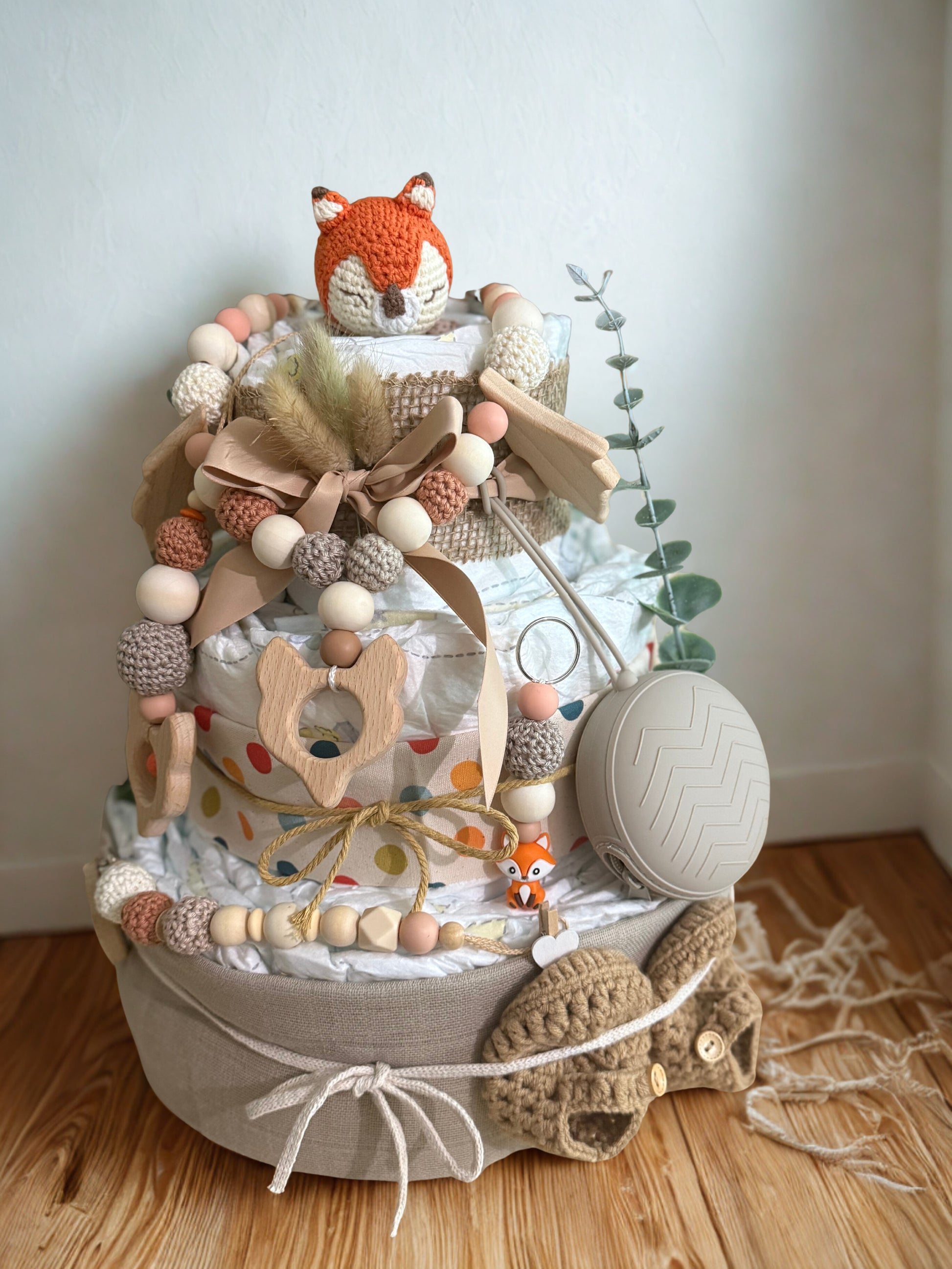 Woodland fox diaper cake baby shower centerpiece featuring three tiers of rolled white diapers wrapped in neutral linen and burlap fabric. Top tier decorated with a crocheted orange fox plush, accented with pampas grass and soft beige ribbon bow. Adorned with wooden and silicone beaded pacifier clip in earthy tones, wooden fox-shaped teething ring, pastel bead garland, and small fox charm detail. Includes beige pacifier case attached to the side and cozy knitted baby booties at the base. Rustic boho newborn