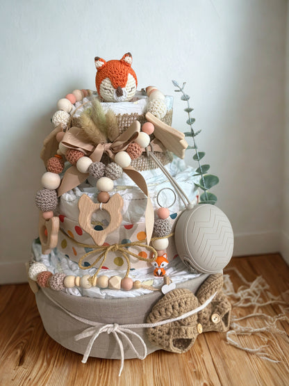 Woodland fox diaper cake baby shower centerpiece featuring three tiers of rolled white diapers wrapped in neutral linen and burlap fabric. Top tier decorated with a crocheted orange fox plush, accented with pampas grass and soft beige ribbon bow. Adorned with wooden and silicone beaded pacifier clip in earthy tones, wooden fox-shaped teething ring, pastel bead garland, and small fox charm detail. Includes beige pacifier case attached to the side and cozy knitted baby booties at the base. Rustic boho newborn