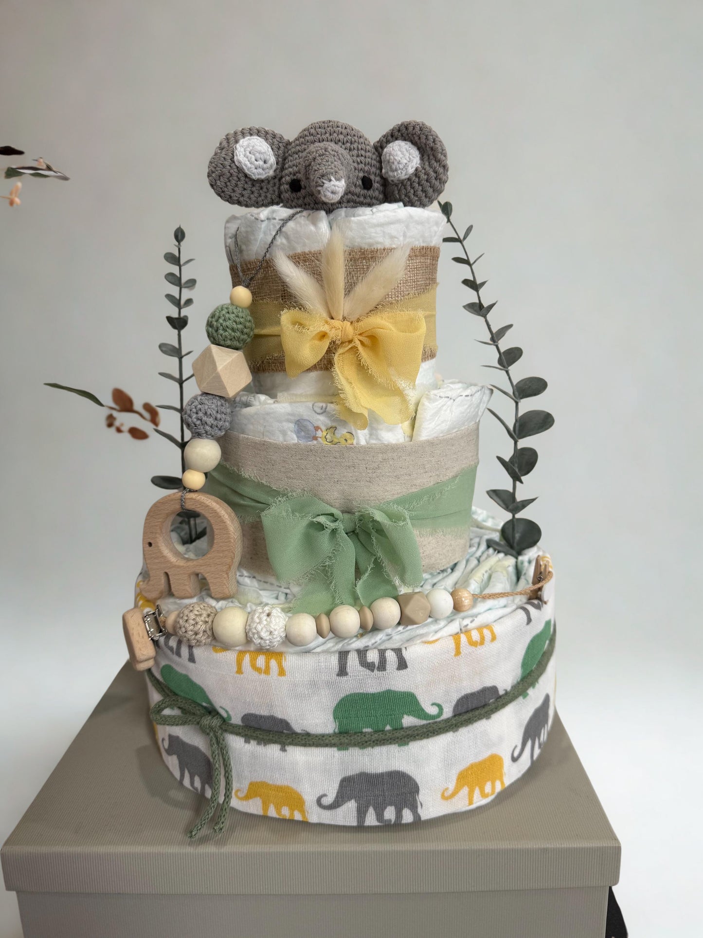 Safari elephant diaper cake baby shower centerpiece featuring three tiers of rolled white diapers wrapped in burlap ribbon and soft sage green fabric bows. Top tier decorated with a crocheted gray elephant plush, accented with dried bunny tail grass and eucalyptus greenery stems. Adorned with wooden elephant-shaped teething ring, silicone and crochet beaded pacifier clip in green, cream, and natural wood tones. Base tier wrapped in white fabric with green, yellow, and gray elephant print, creating a playful