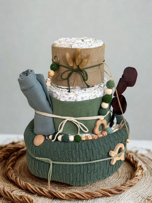 Green and natural woodland diaper cake baby shower gift featuring three tiers of rolled white diapers wrapped in sage green fabric, burlap ribbon, and textured knit basket base. Decorated with wooden and silicone beaded pacifier clip personalized with letter beads spelling “MANNI,” wooden teething ring, star-shaped wooden charm, and earthy green crochet beads. Accented with dried pampas grass, dark green twine bow, and neutral baby blanket rolled on the side. Rustic boho nursery centerpiece styled on a wove