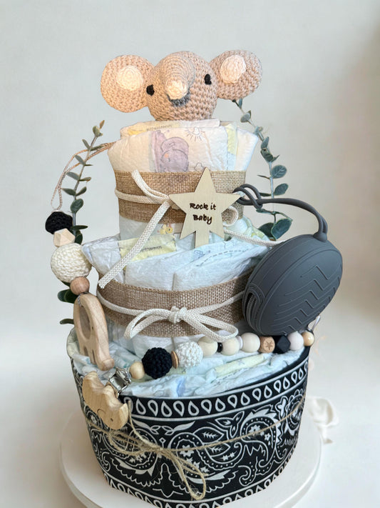 Modern neutral diaper cake baby shower centerpiece featuring three tiers of rolled white diapers wrapped in natural burlap ribbon and soft cream cord bows. Top tier decorated with a crocheted beige elephant plush, accented with eucalyptus greenery stems. Wooden star-shaped tag with engraved text “Rock it Baby” attached to the middle tier for a personalized touch. Adorned with wooden elephant teething ring, silicone and crochet beaded pacifier clip in black, white, and natural wood tones, plus dark gray paci